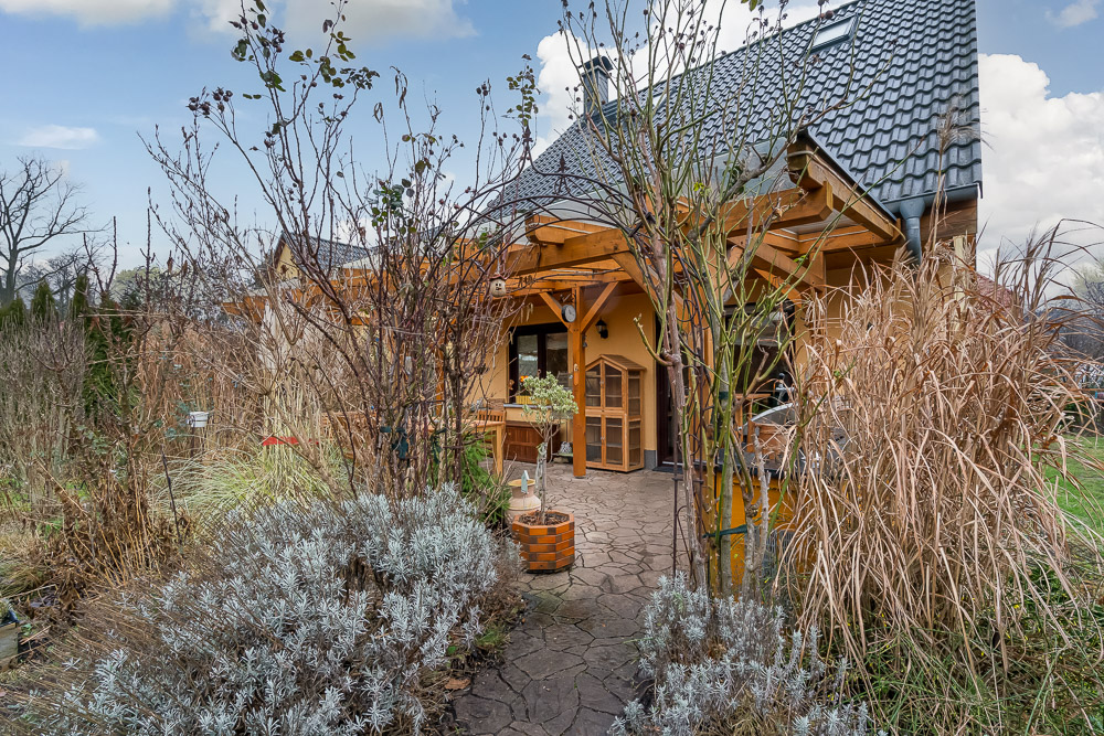 Gartenweg mit Pflanzen führt zu einem Haus mit Holzveranda und dunklem Dach.