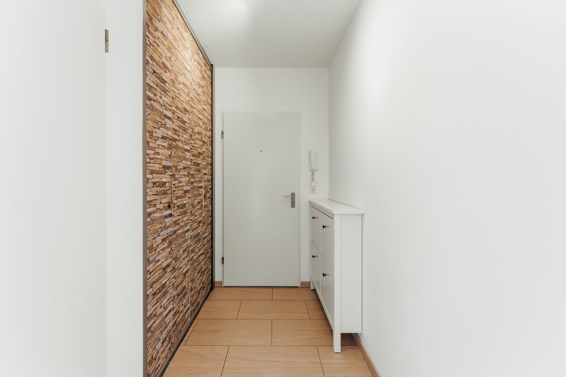 Heller Flur mit Holzboden, weißer Kommode, Tür und Wand mit Steinverkleidung.