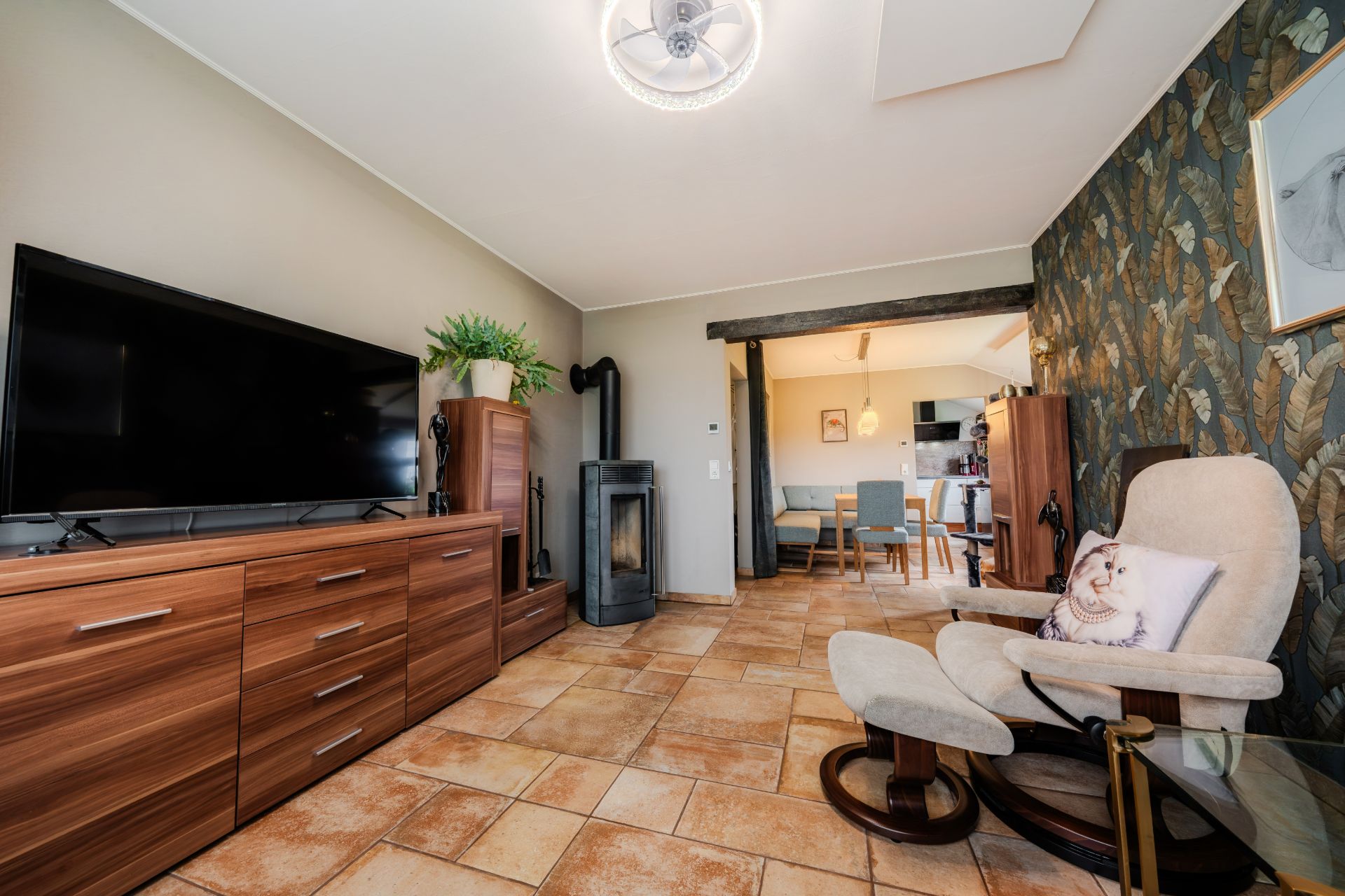 Wohnzimmer mit Fernseher, Holzschränken, Sessel, Kaminofen und Essbereich im Hintergrund.