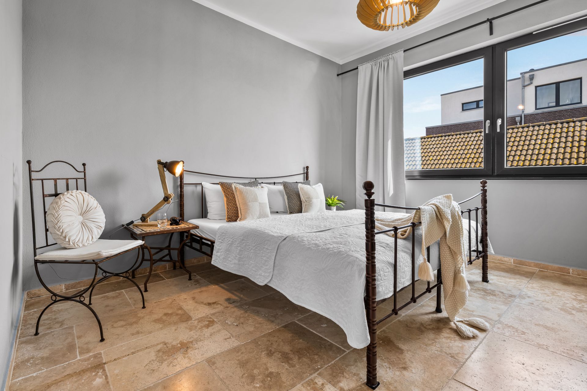 Helles Schlafzimmer mit Doppelbett, Eisenrahmen, Stuhl, Nachttischlampe und großem Fenster mit Blick nach draußen.