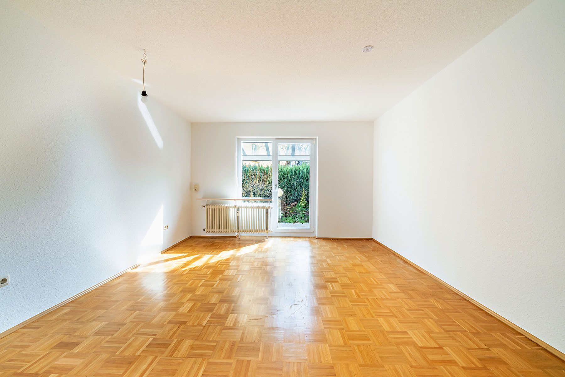 Heller, leerer Raum mit Parkettboden, weißen Wänden und Fenstertür zum Garten.