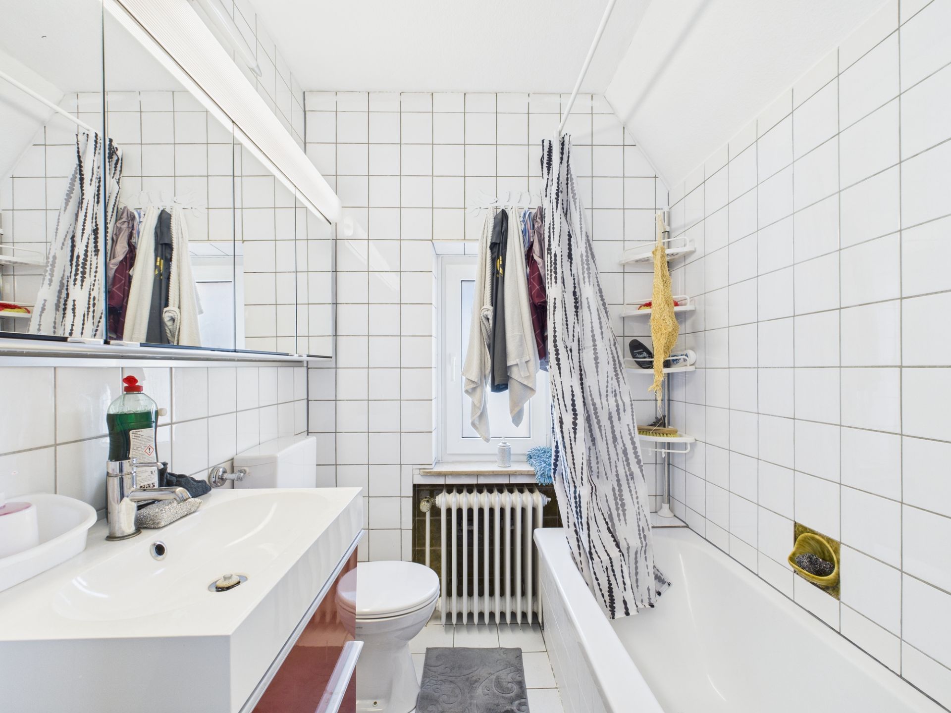 Helles Badezimmer mit Badewanne, Waschbecken, Spiegel und Duschvorhang, weiße Fliesen an den Wänden.