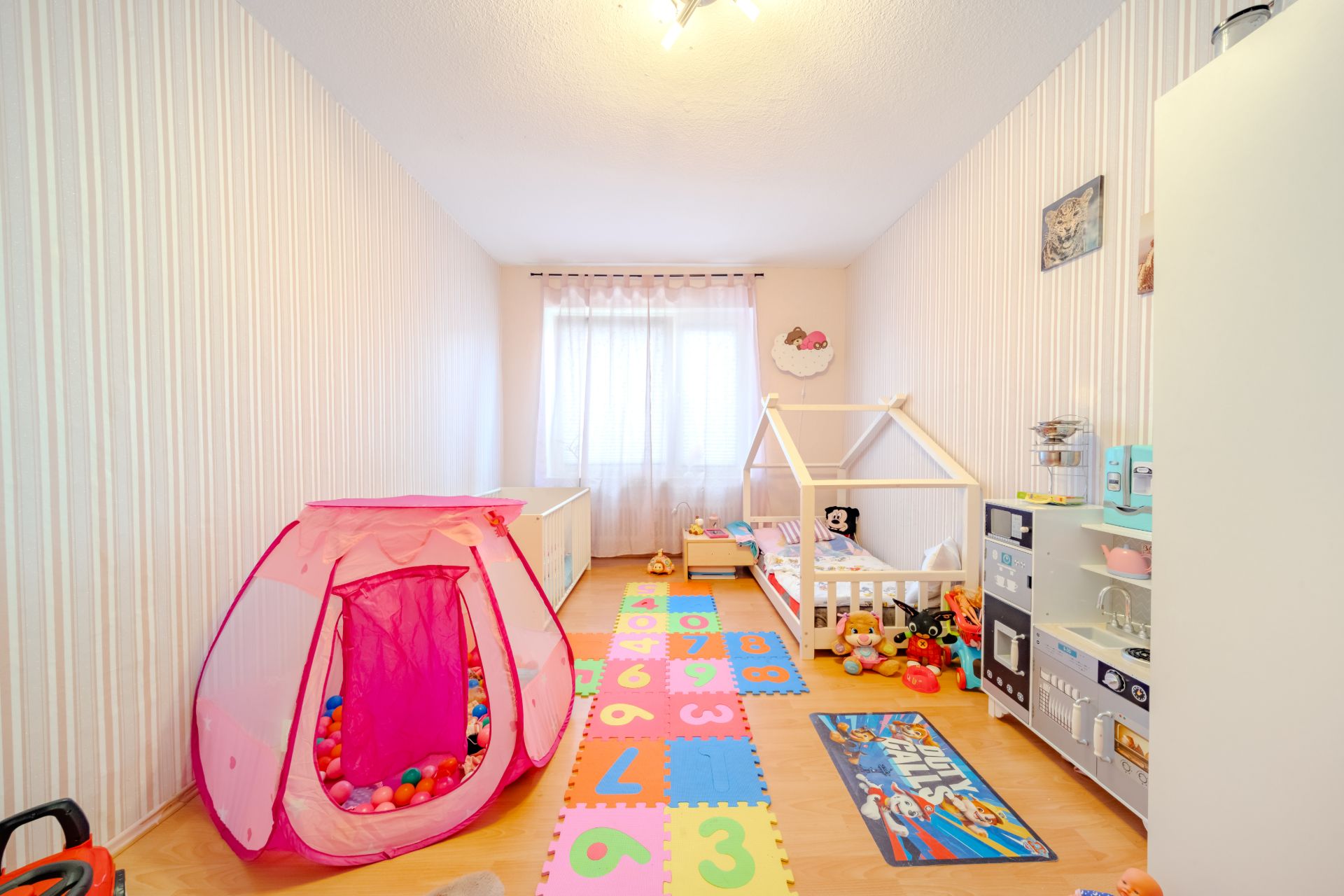 Helles Kinderzimmer mit Spielzelt, Spielzeugküche, Teppich mit Zahlen und Kinderbett.
