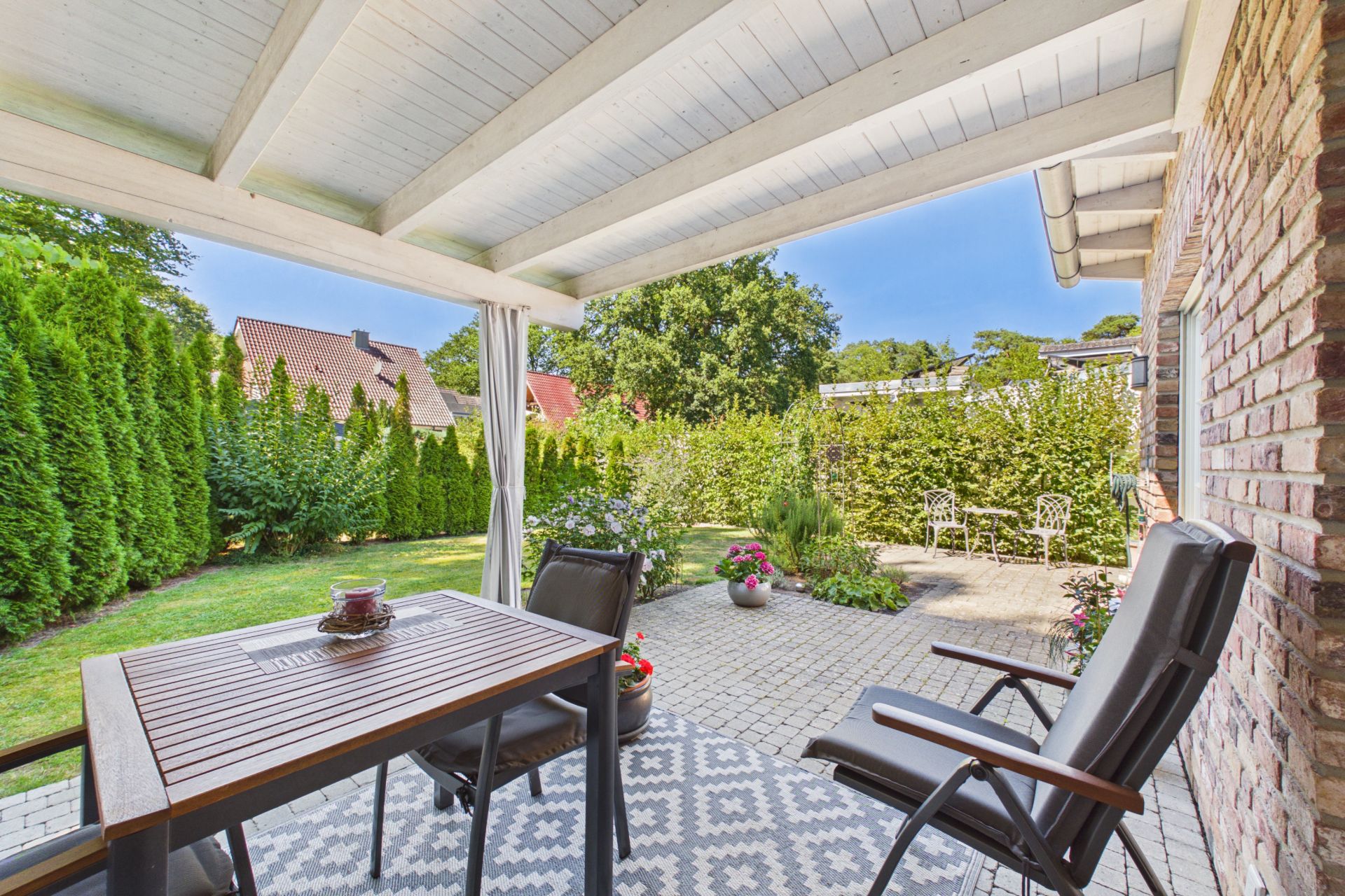 Überdachte Terrasse mit Tisch, Stühlen und Blick auf Garten mit Pflanzen und Sitzbereich.