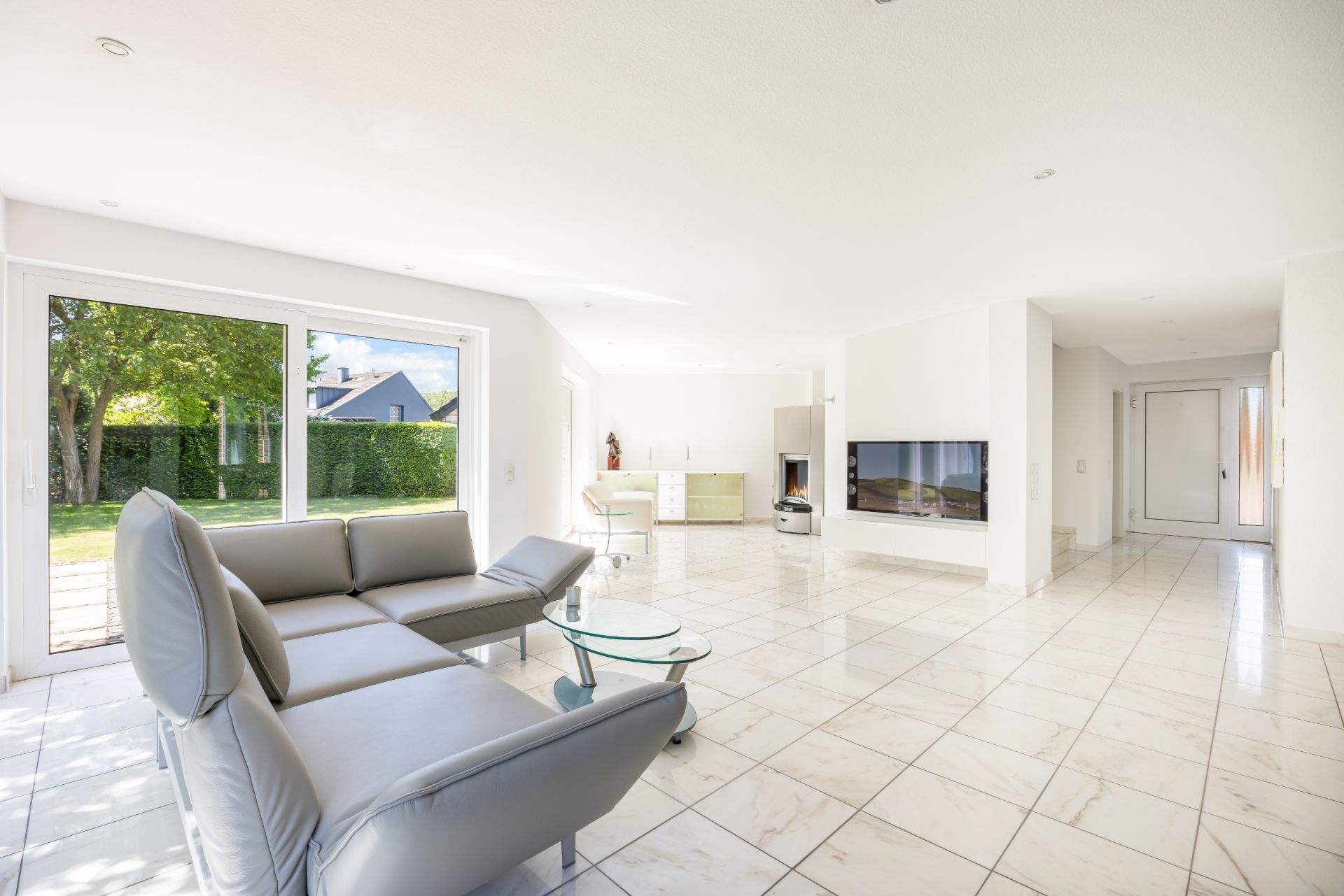 Helles Wohnzimmer mit grauem Sofa, Glastisch und Fliesenboden, große Fenster mit Blick in den Garten.