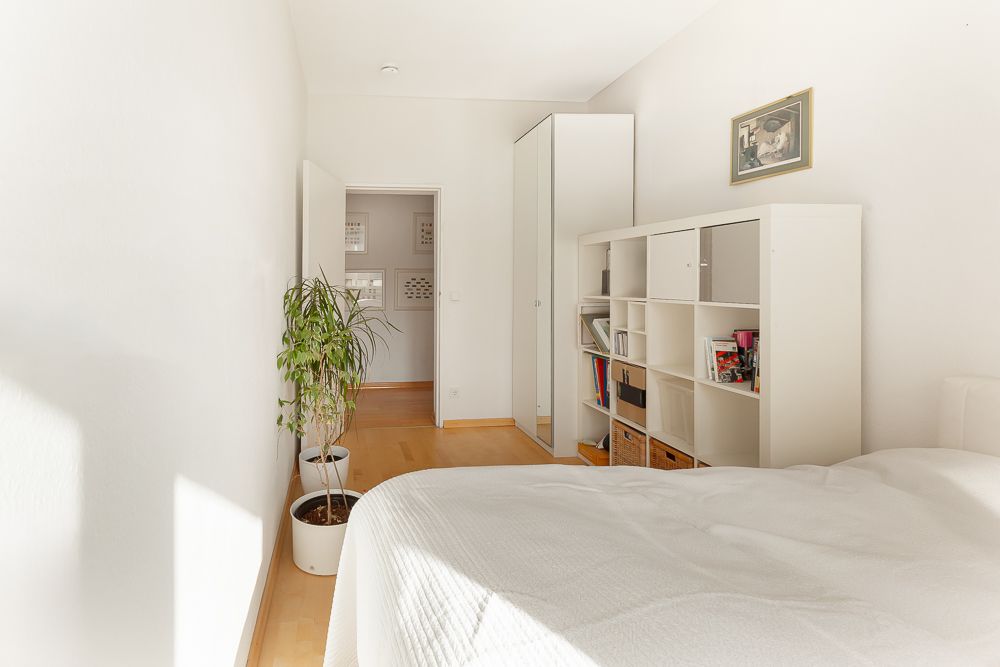 Helles Schlafzimmer mit Bett, Regal, Schrank und Zimmerpflanze auf Holzfußboden.