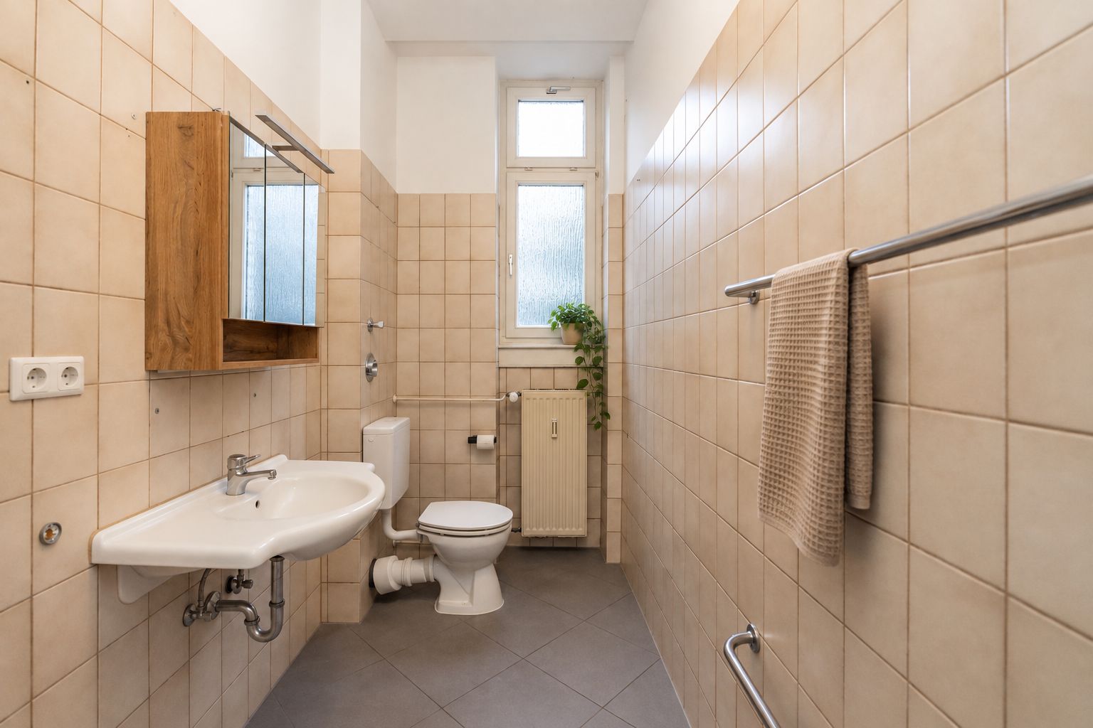 Helles Badezimmer mit Waschbecken, Toilette, Handtuch und Spiegel, Fliesenwänden und Fenster mit Pflanze.