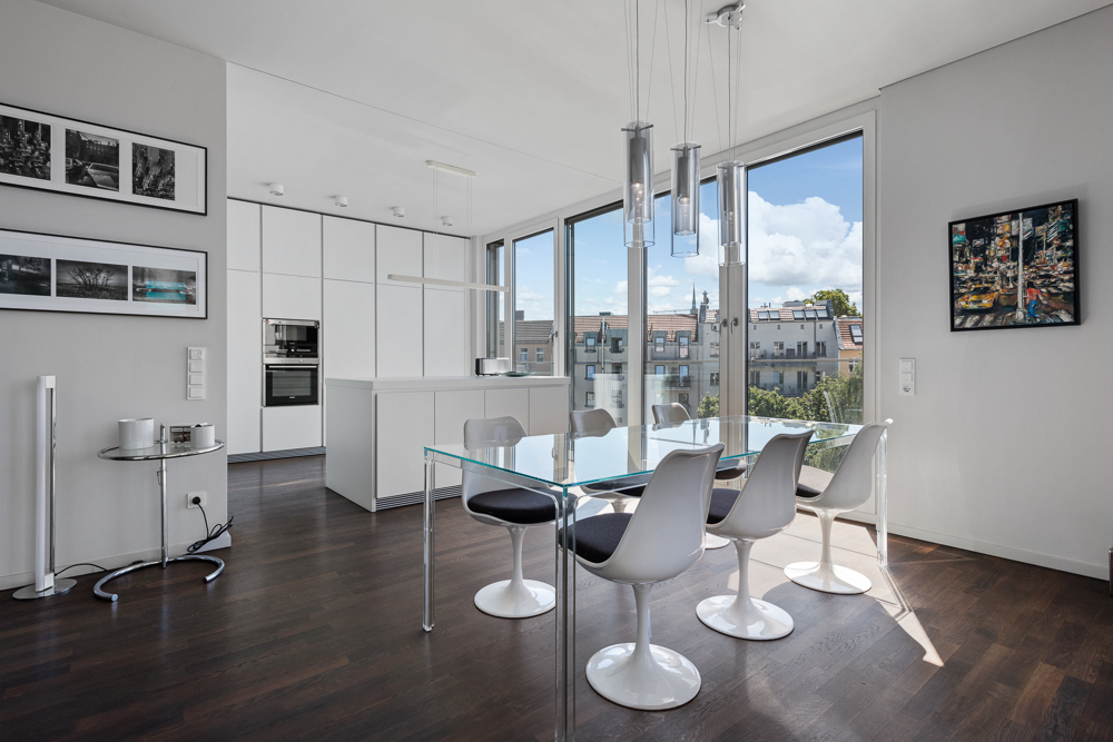 Heller Essbereich mit Glastisch, sechs weißen Stühlen und großer Fensterfront zur Stadt.