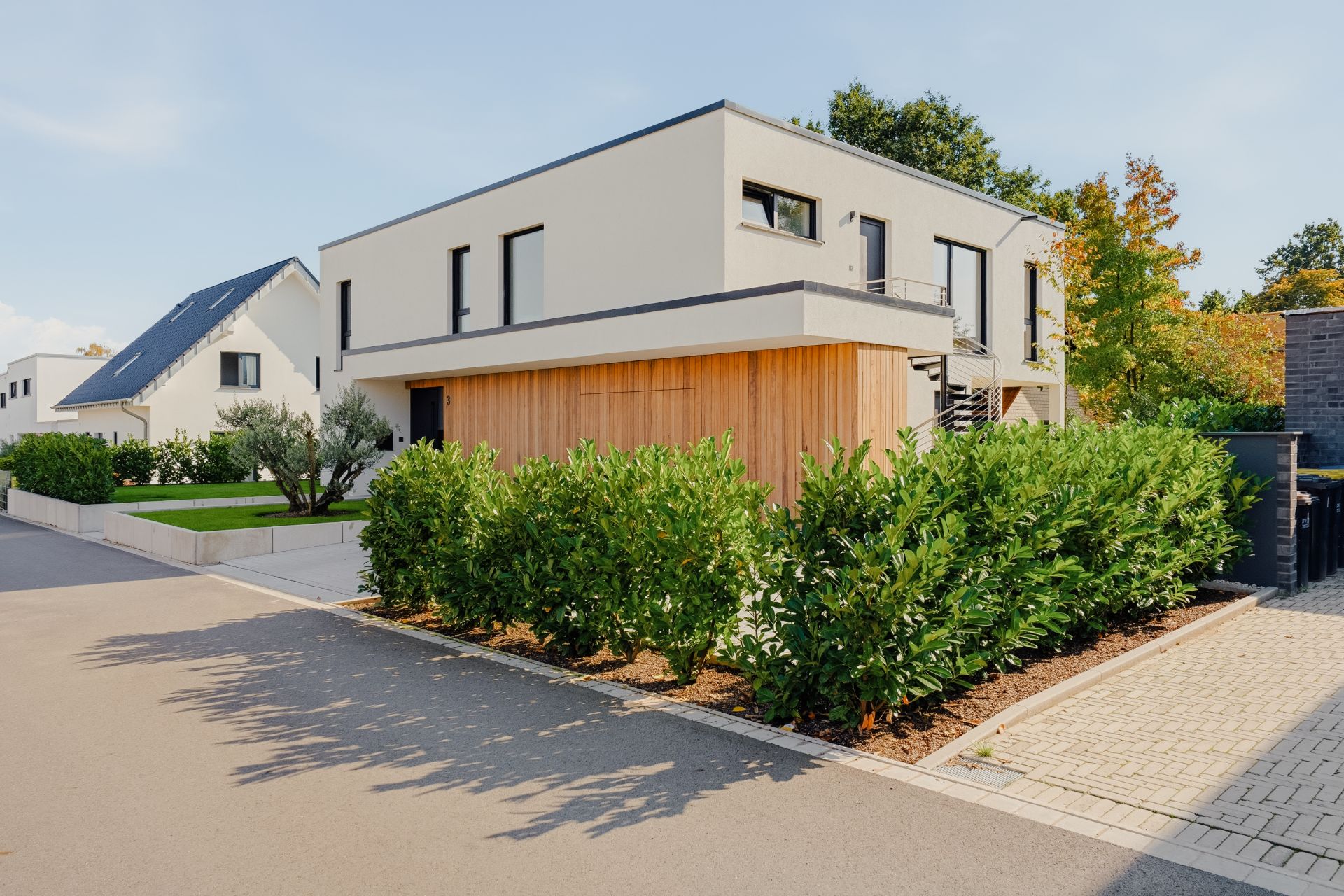Modernes zweistöckiges Wohnhaus mit Flachdach, Holzfassade und grünen Sträuchern im Vorgarten.