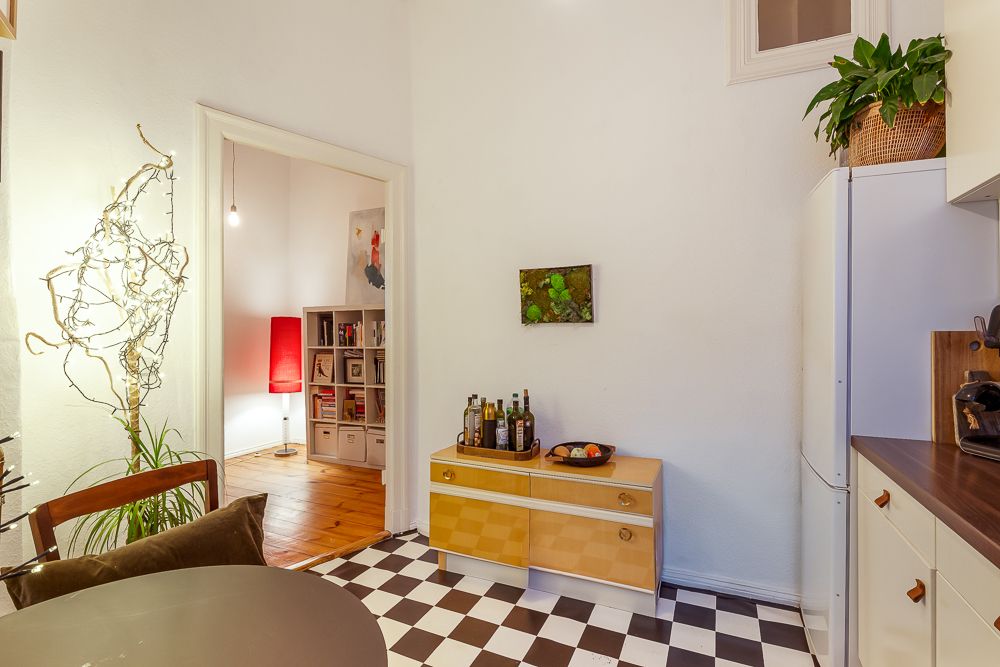 Helle Küche mit Schachbrettboden, Pflanzen, Kommode und Blick ins Wohnzimmer mit roter Stehlampe.