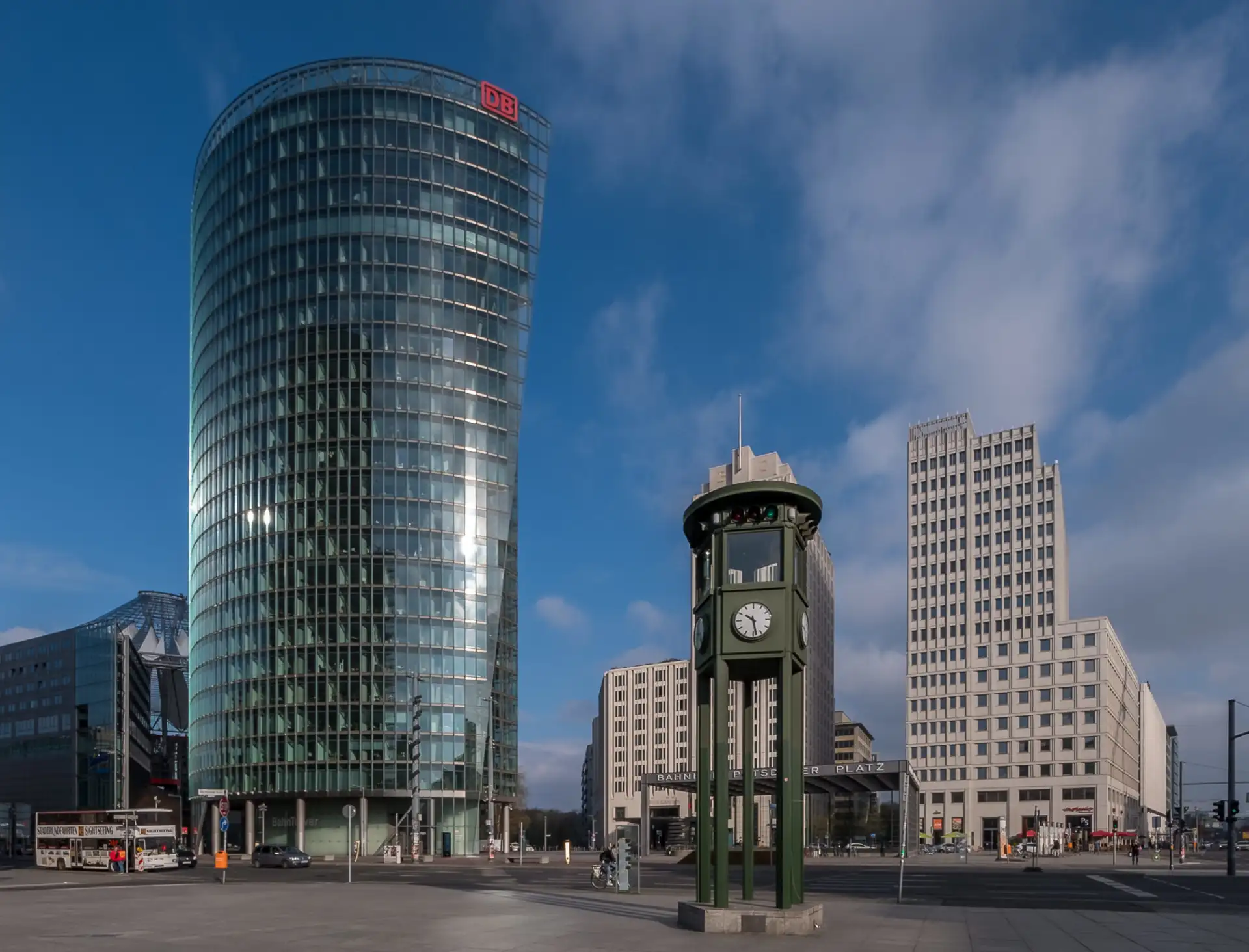 Städtischer Platz mit modernen Hochhäusern und einer großen Uhr auf einem Sockel.