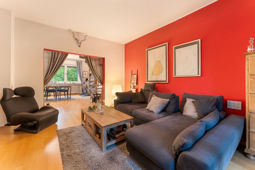 Wohnzimmer mit roter Wand, grauem Sofa, Sessel und Blick in Essbereich mit großem Fenster.