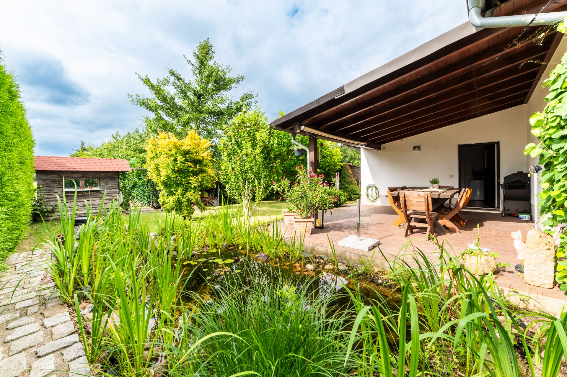 Garten mit Teich, Terrasse mit Holzdach und Sitzgruppe, umgeben von Bäumen und Pflanzen.