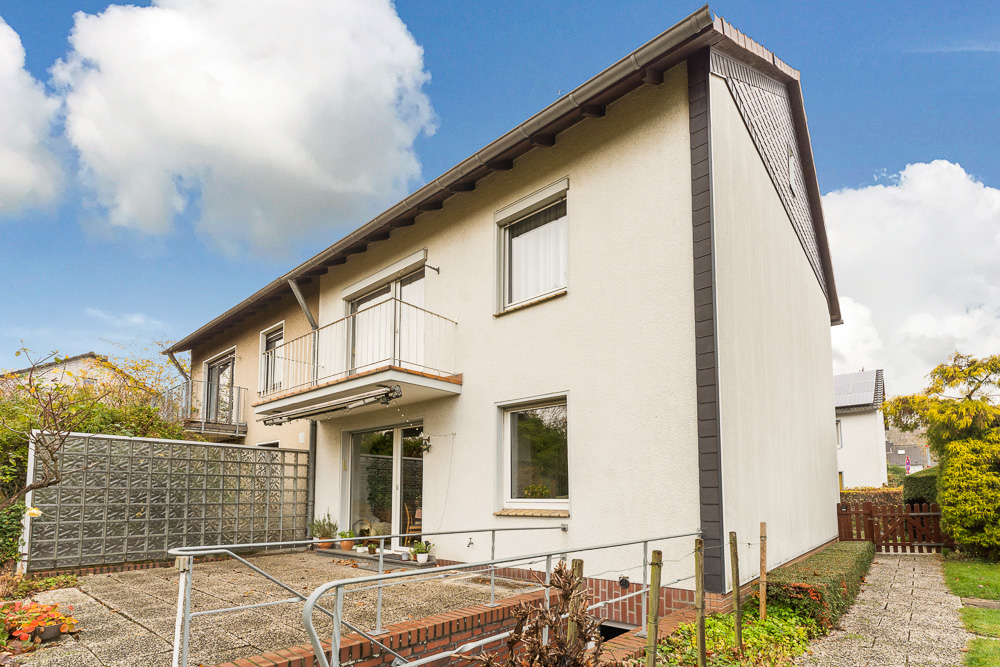 Zweistöckiges Wohnhaus mit Balkon und Terrasse, umgeben von Garten und gepflasterten Wegen.