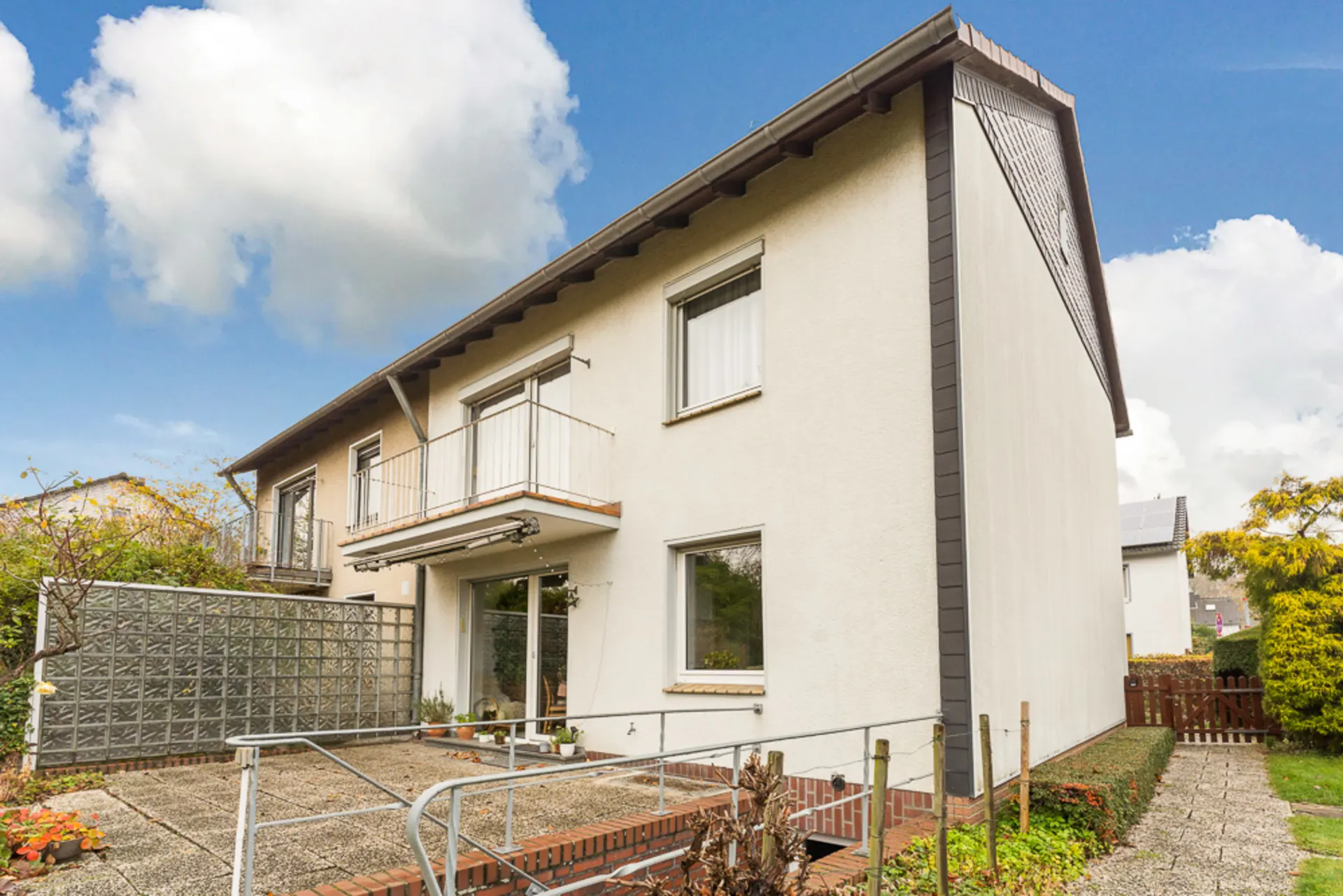 Zweistöckiges Wohnhaus mit Balkon und Terrasse, umgeben von Garten und gepflasterten Wegen.