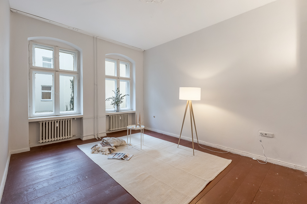 Heller Raum mit zwei Fenstern, Stehlampe, Teppich und kleinem Tisch auf Holzboden.