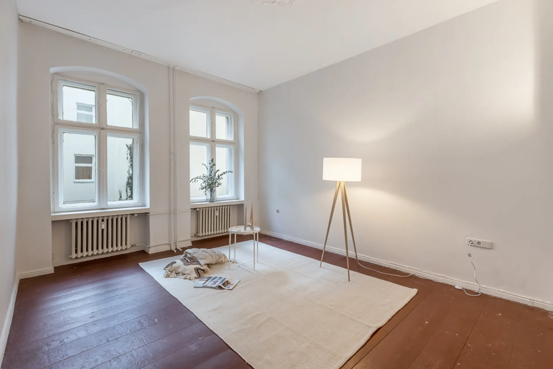 Heller Raum mit zwei Fenstern, Stehlampe, Teppich und kleinem Tisch auf Holzboden.
