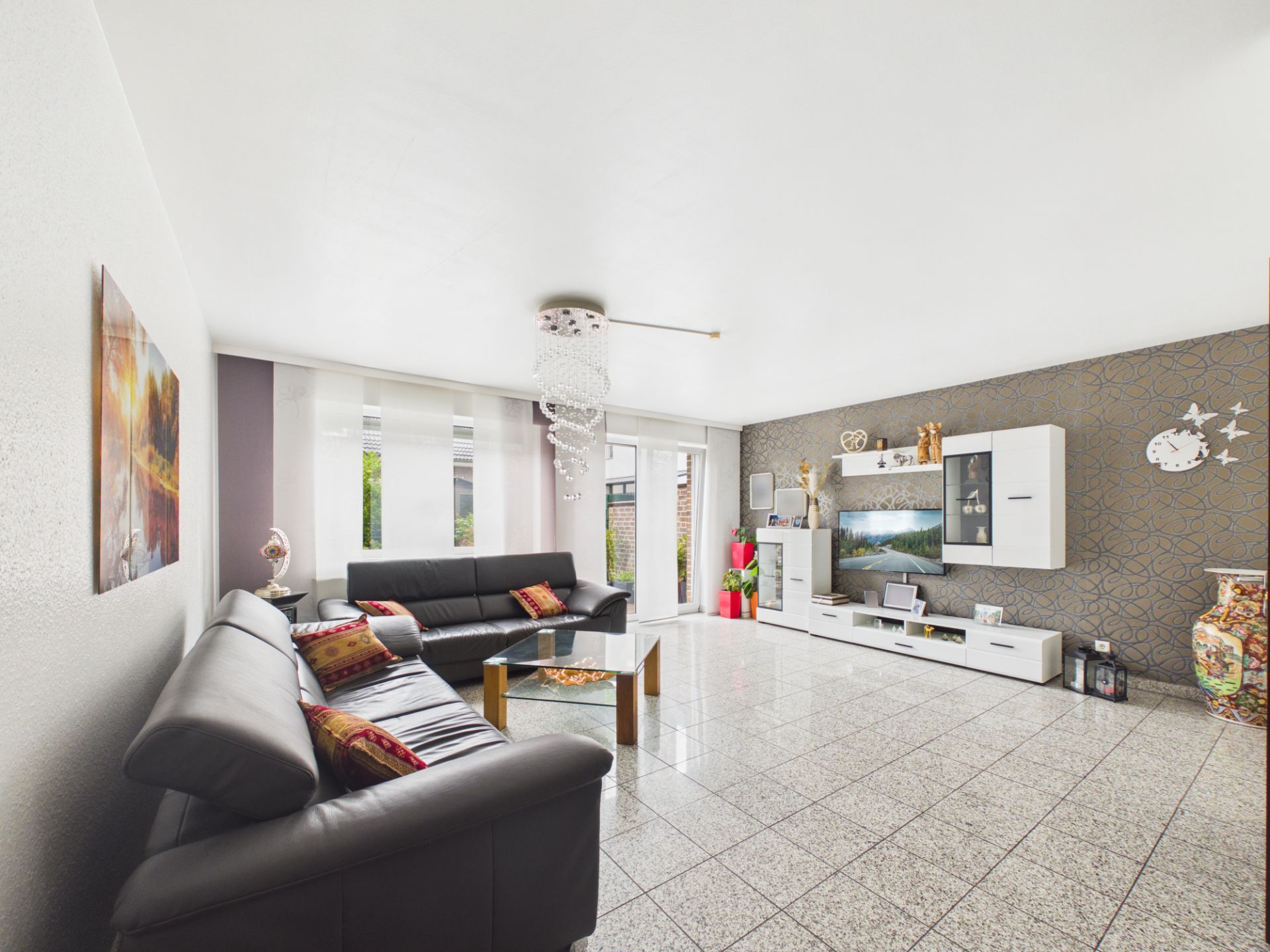Modernes Wohnzimmer mit zwei Sofas, Glastisch und TV-Wand, große Fenster lassen viel Licht herein.