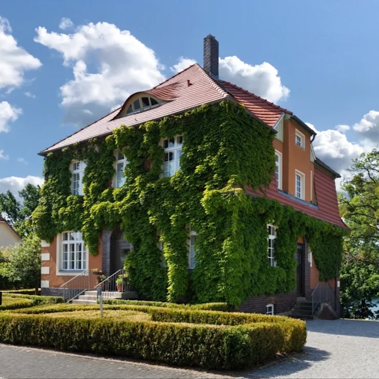 Großes Wohnhaus mit rotem Dach, Fassadenbegrünung und gepflegtem Vorgarten bei sonnigem Wetter.