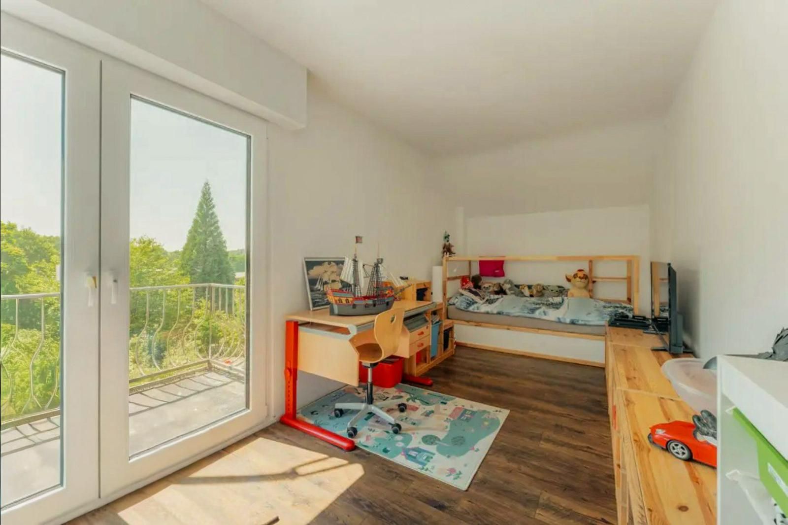 Helles Kinderzimmer mit Schreibtisch, Bett, großem Fenster und Blick ins Grüne.