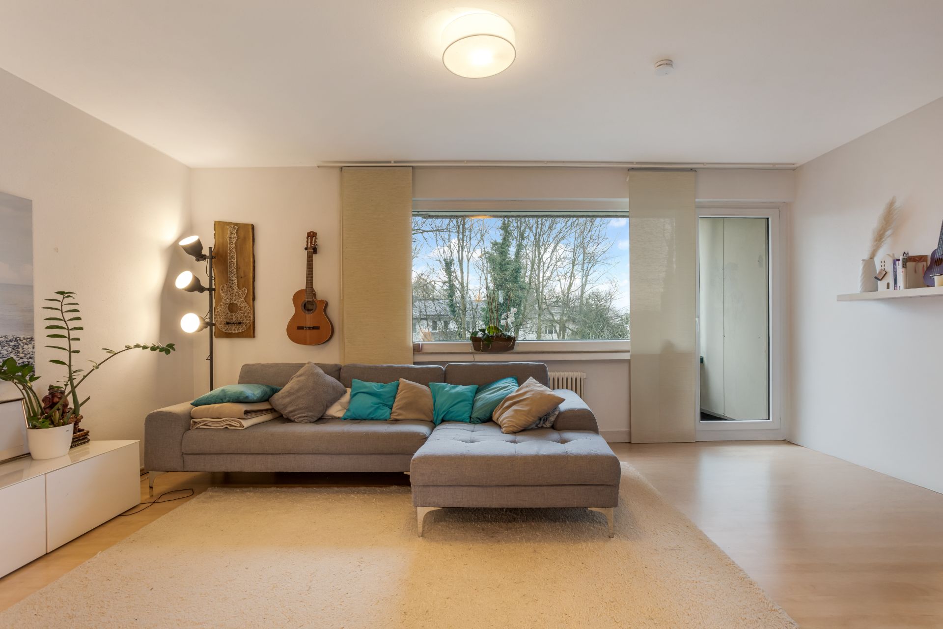 Helles Wohnzimmer mit grauem Sofa, großen Fenstern und dekorativer Gitarre an der Wand.