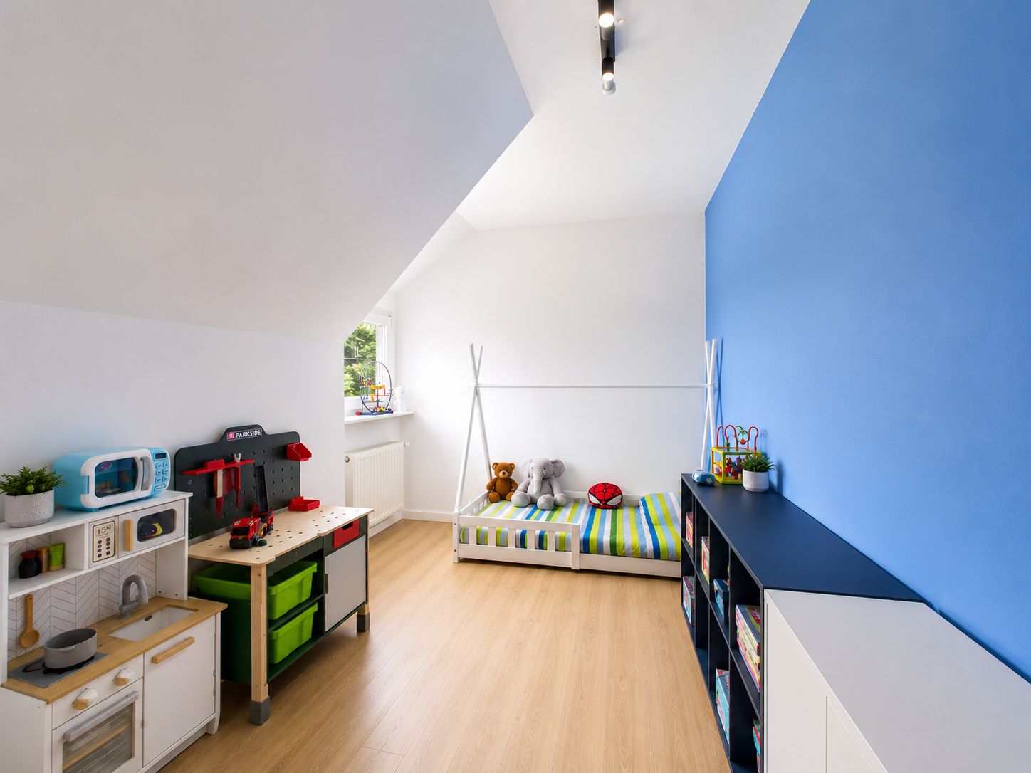 Kinderzimmer mit Bett, Spielküche, Spielsachen und Regal. Eine Wand ist blau gestrichen.