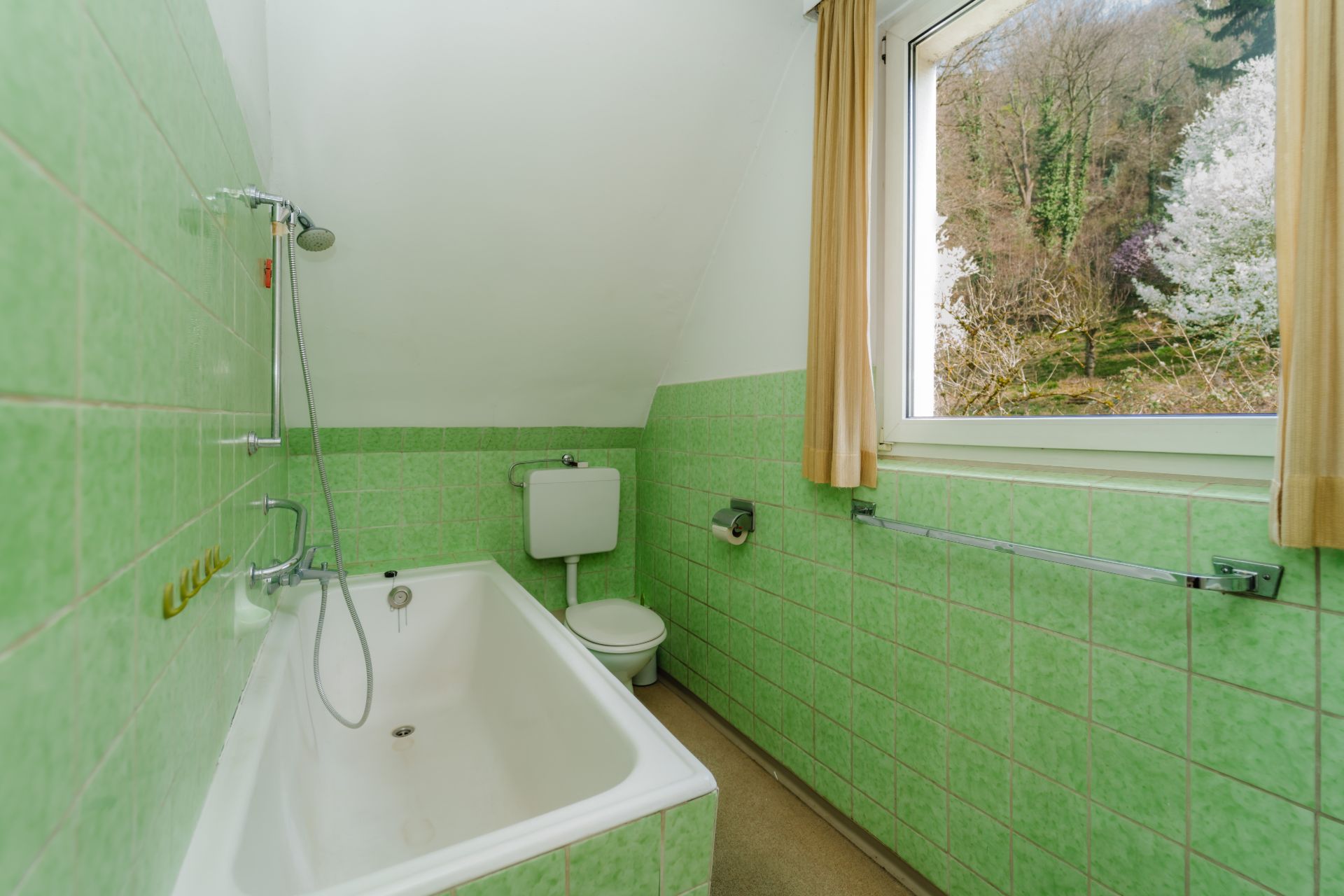 Badezimmer mit grünen Fliesen, Badewanne, Toilette und Fenster mit Blick nach draußen.