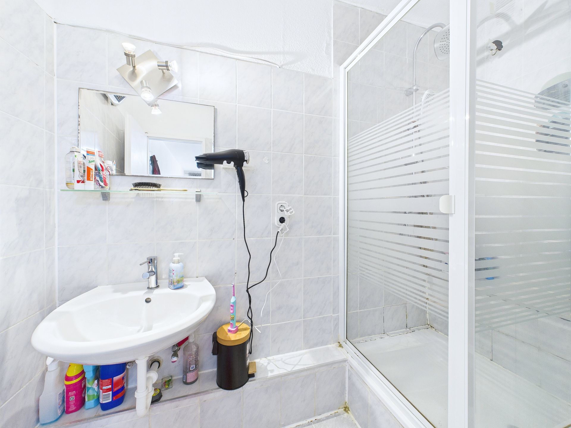 Kleines Badezimmer mit Waschbecken, Spiegel, Duschkabine und Hygieneartikeln.