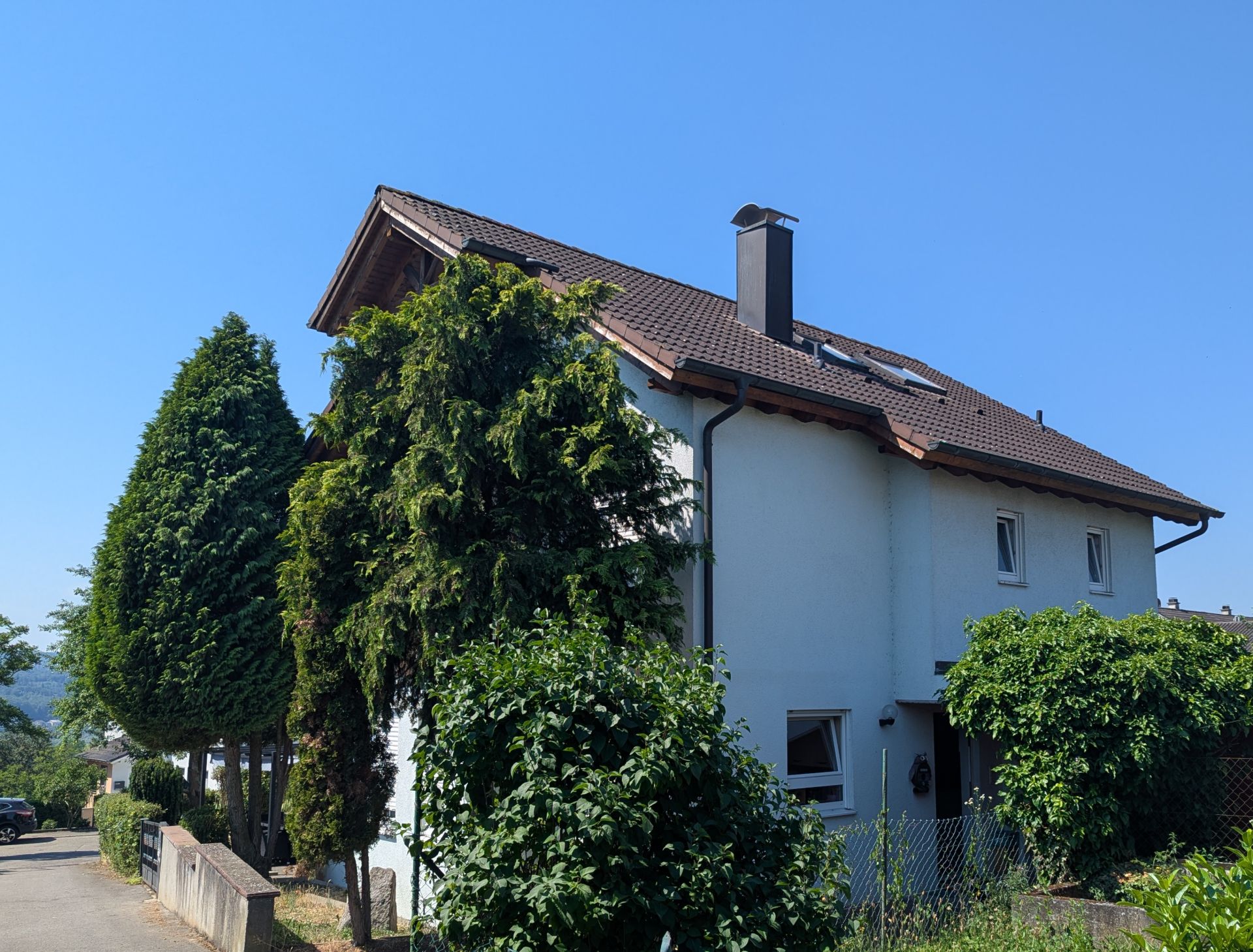 Zweistöckiges Wohnhaus mit braunem Dach, umgeben von Bäumen und Sträuchern an einer ruhigen Straße.