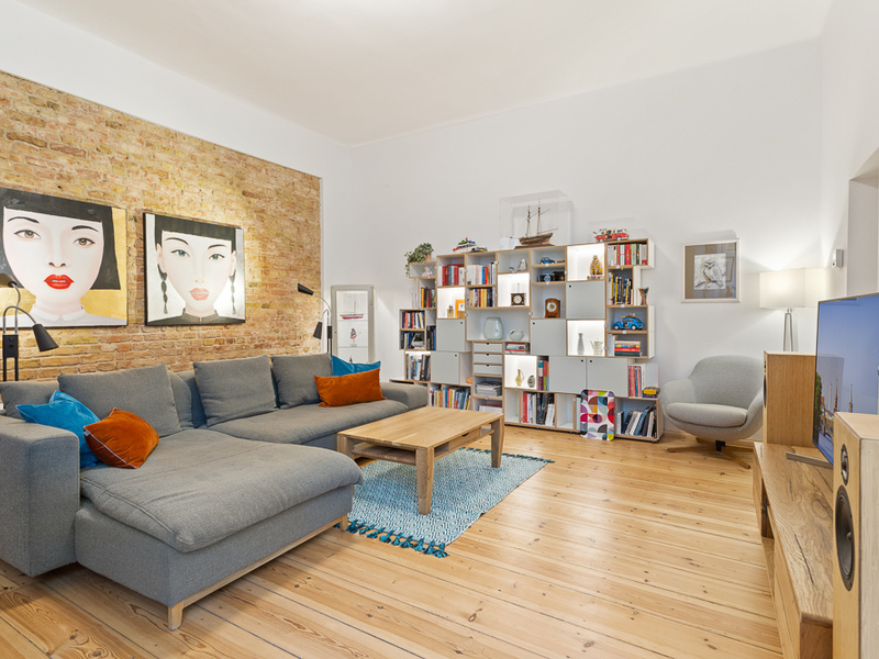 Modernes Wohnzimmer mit grauem Sofa, Regalwand, Holztisch und Bildern an einer Backsteinwand.