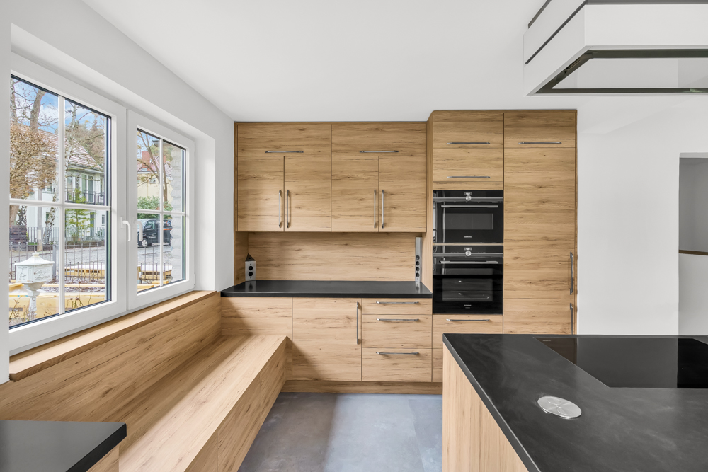 Moderne Küche mit Holzfronten, schwarzer Arbeitsplatte, Sitzbank und großen Fenstern.