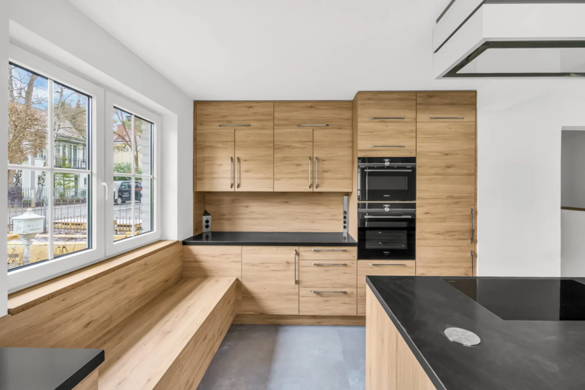 Moderne Küche mit Holzfronten, schwarzer Arbeitsplatte, Sitzbank und großen Fenstern.