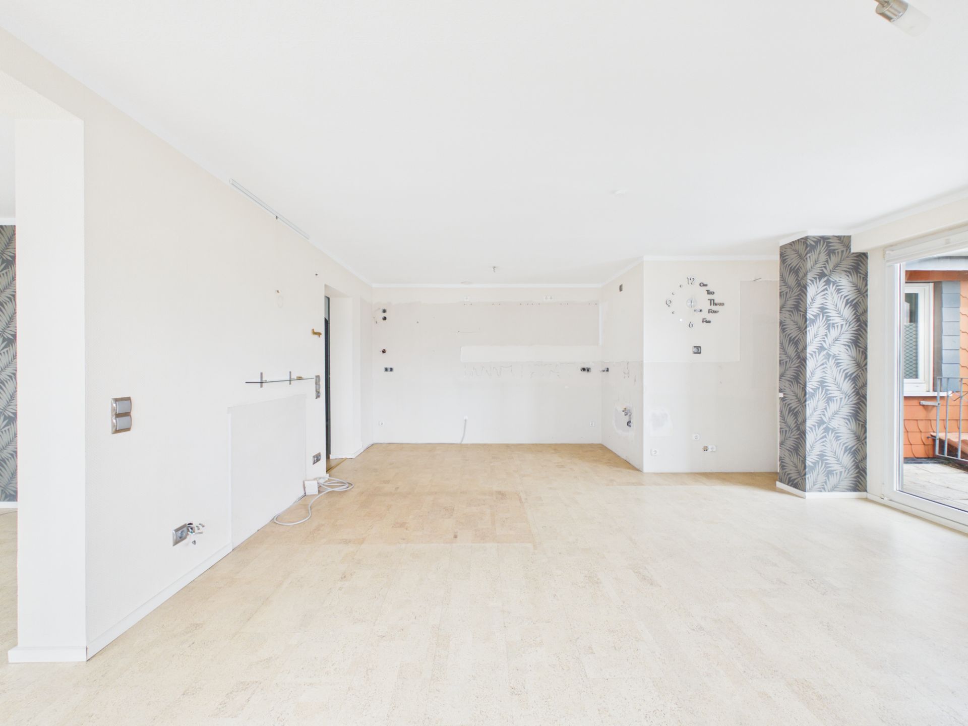 Leerer, heller Raum mit Holzfußboden und großen Fenstern, Wand mit Tapetenstreifen und Balkonzugang.