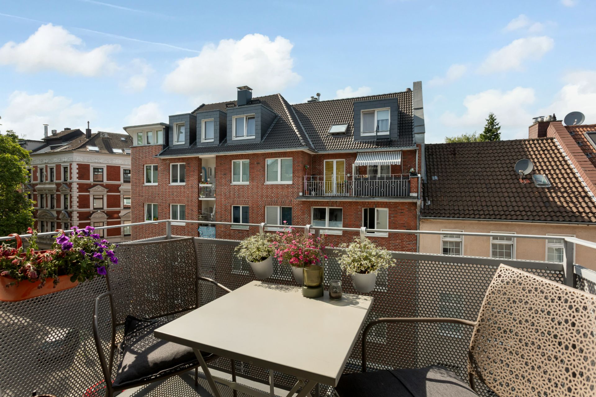Balkon mit Tisch, Stühlen und Blumen, Ausblick auf benachbarte Wohngebäude.