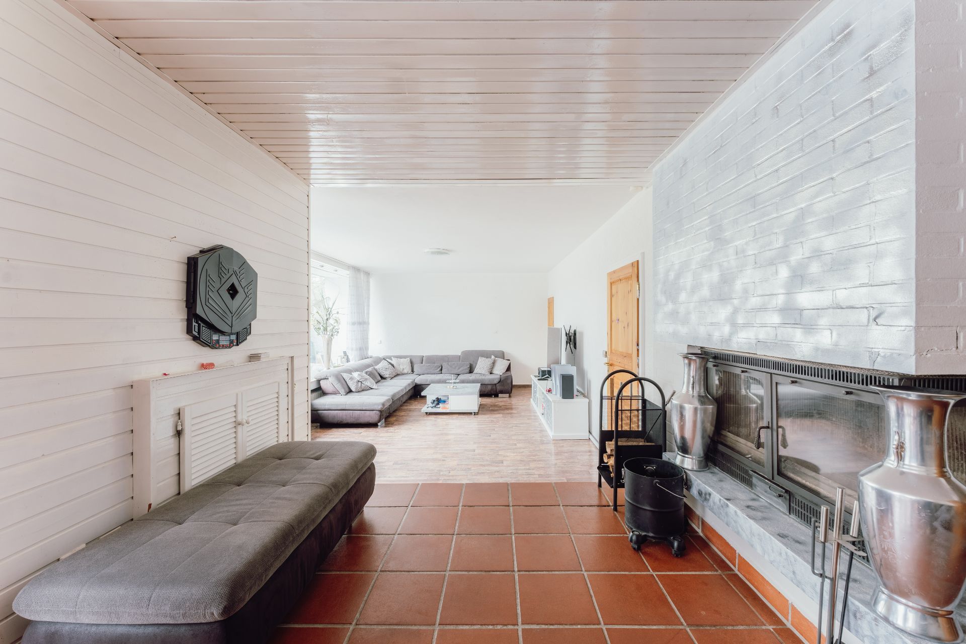 Helles Wohnzimmer mit Ecksofa, Kamin, grauer Bank und großen Fenstern.