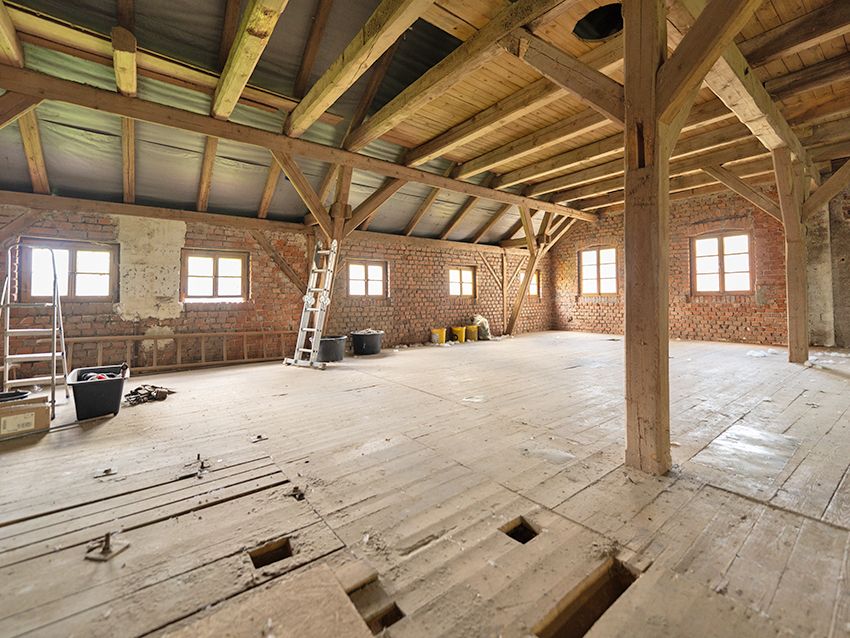 Ausgebauter Dachboden mit Holzbalken, Holzboden, Leitern und Fenstern, teils mit Baumaterialien.