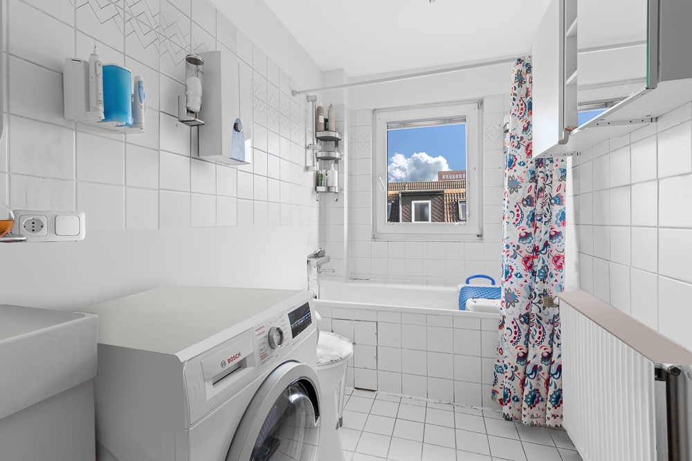 Helles Badezimmer mit Waschmaschine, Badewanne, Fenster und gemusterter Duschvorhang.