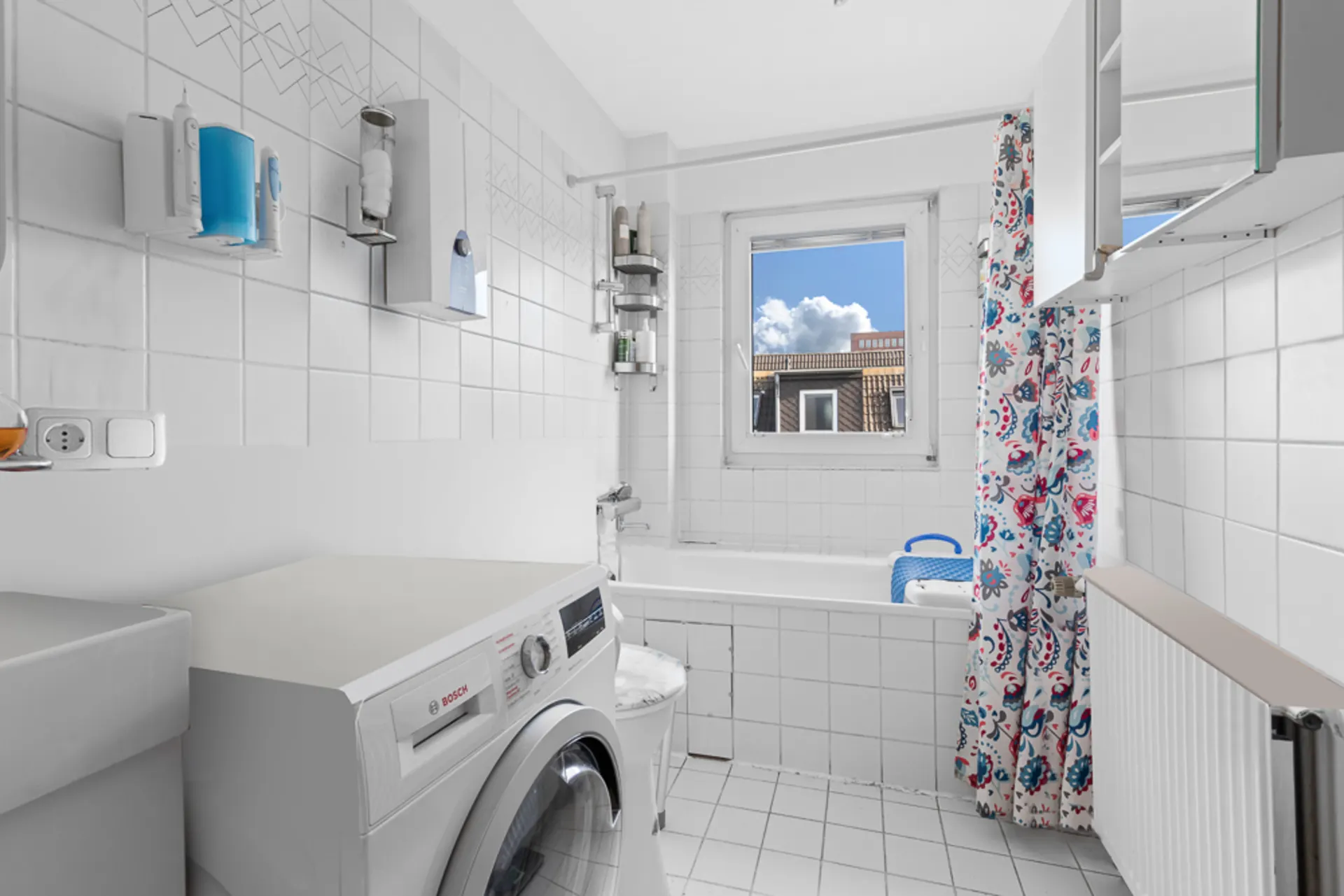 Helles Badezimmer mit Waschmaschine, Badewanne, Fenster und gemusterter Duschvorhang.