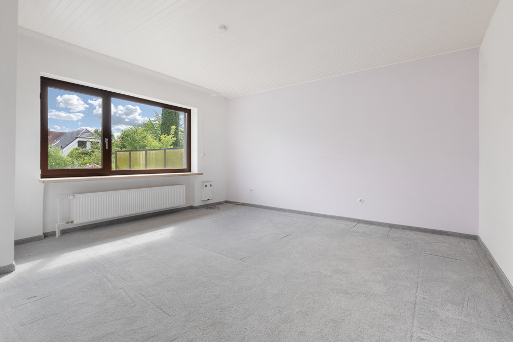 Helles, leeres Zimmer mit großem Fenster, Teppichboden und weiß gestrichenen Wänden.