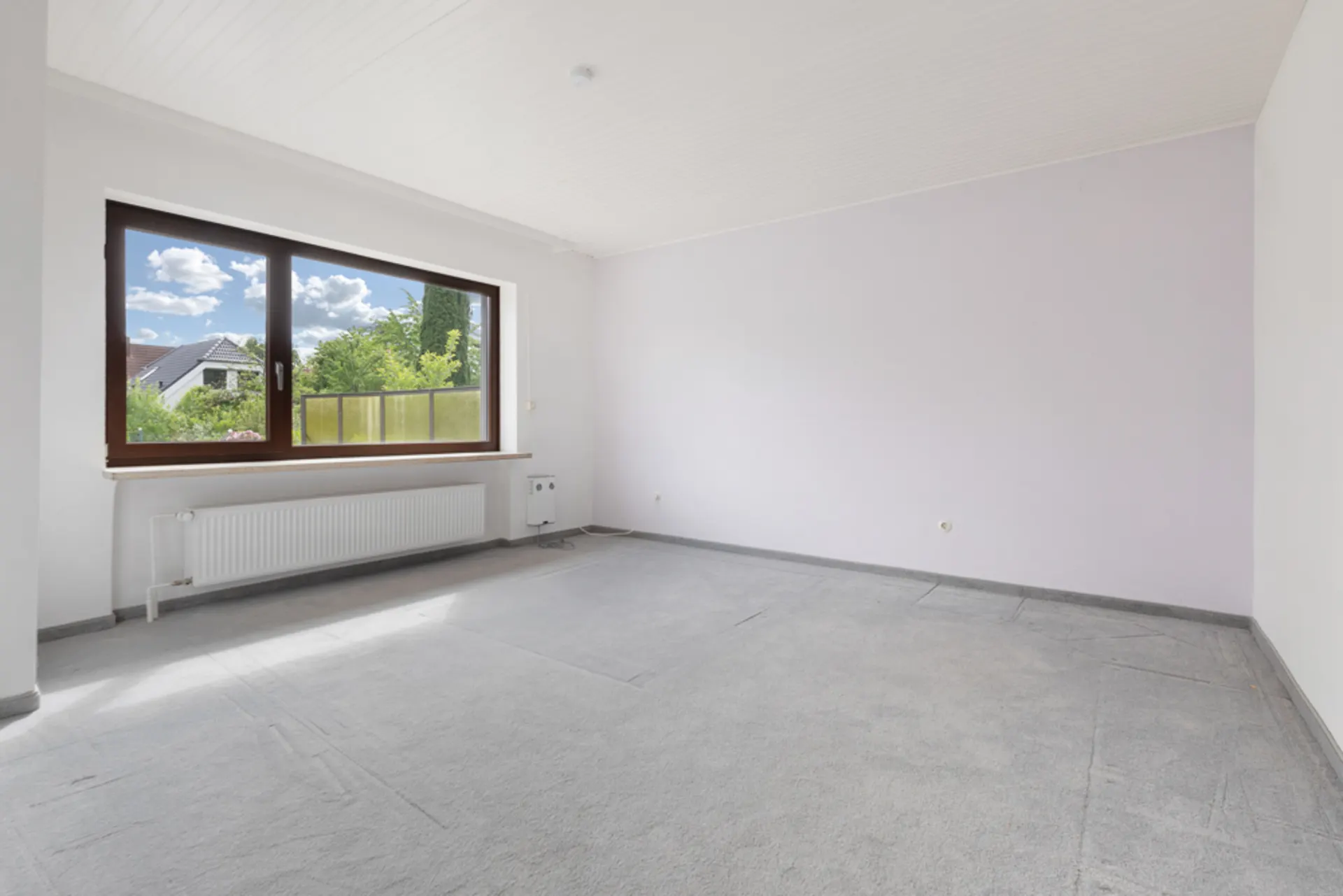 Helles, leeres Zimmer mit großem Fenster, Teppichboden und weiß gestrichenen Wänden.