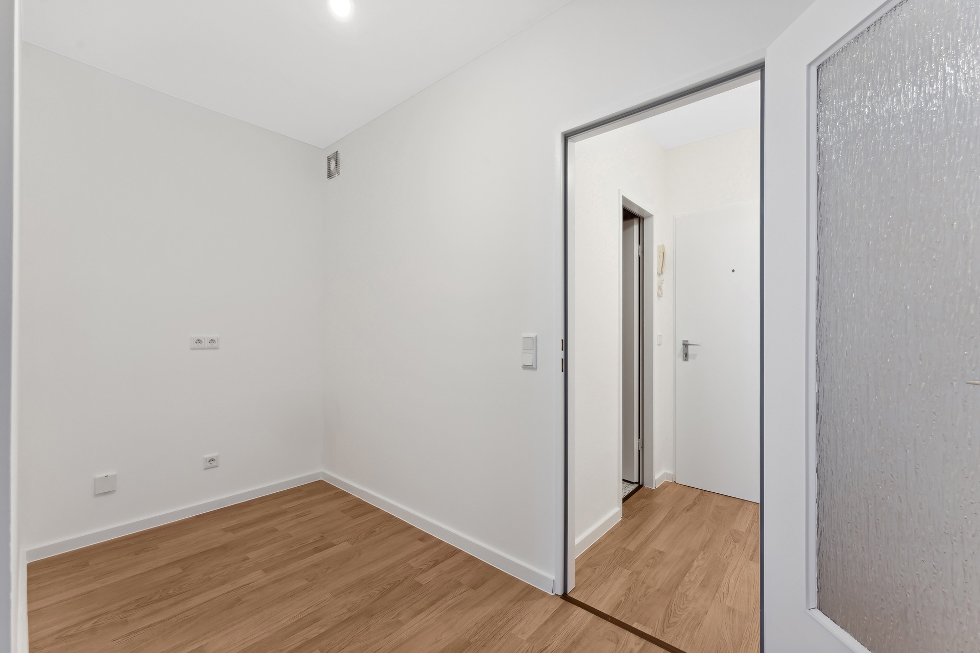 Leerer Raum mit weißer Wand, Holzboden und Tür zu einem Flur.