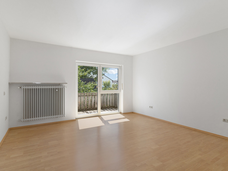 Helles, leeres Zimmer mit Holzfußboden, großem Fenster und Balkonzugang.