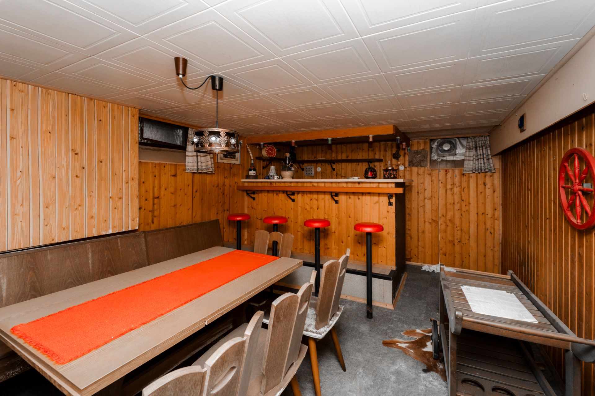 Rustikaler Raum mit Holzwänden, langer Esstisch, Bar mit roten Hockern, dekorativer Leuchte.