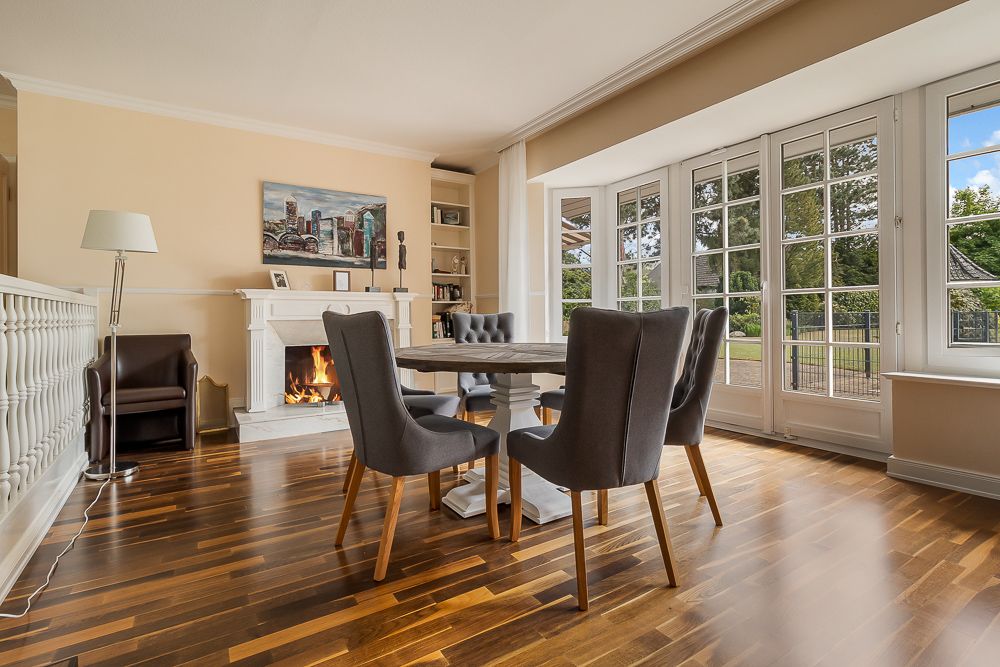 Heller Wohnraum mit Kamin, rundem Tisch und sechs Stühlen, große Fenster mit Blick nach draußen.