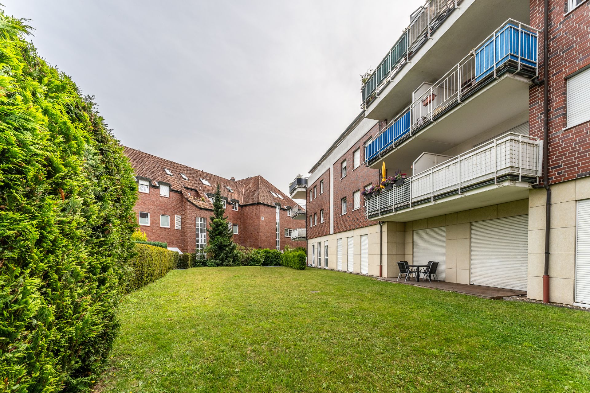 Grüne Wiese mit Hecke, davor ein Mehrfamilienhaus mit Balkonen und Terrasse.
