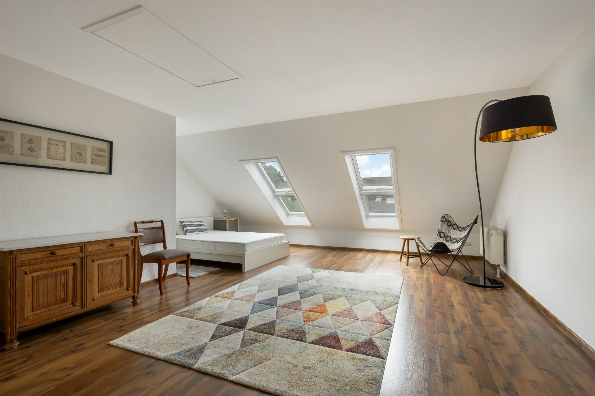 Helles Schlafzimmer mit Holzboden, Teppich, Bett, Kommode, Stuhl und zwei Dachfenstern.