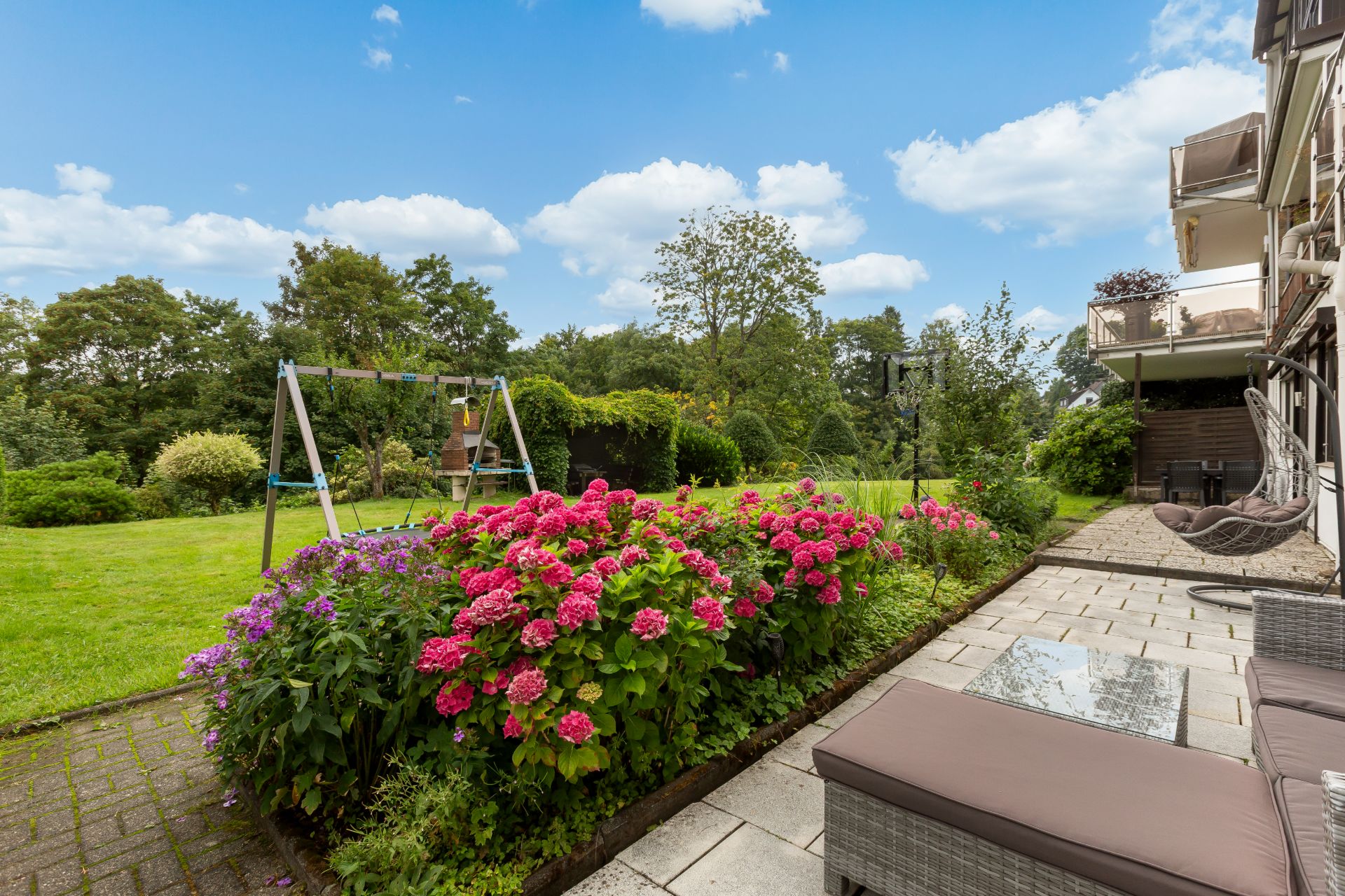 Garten mit Blumenbeet, Terrasse mit Sitzmöbeln und Schaukel im Hintergrund.