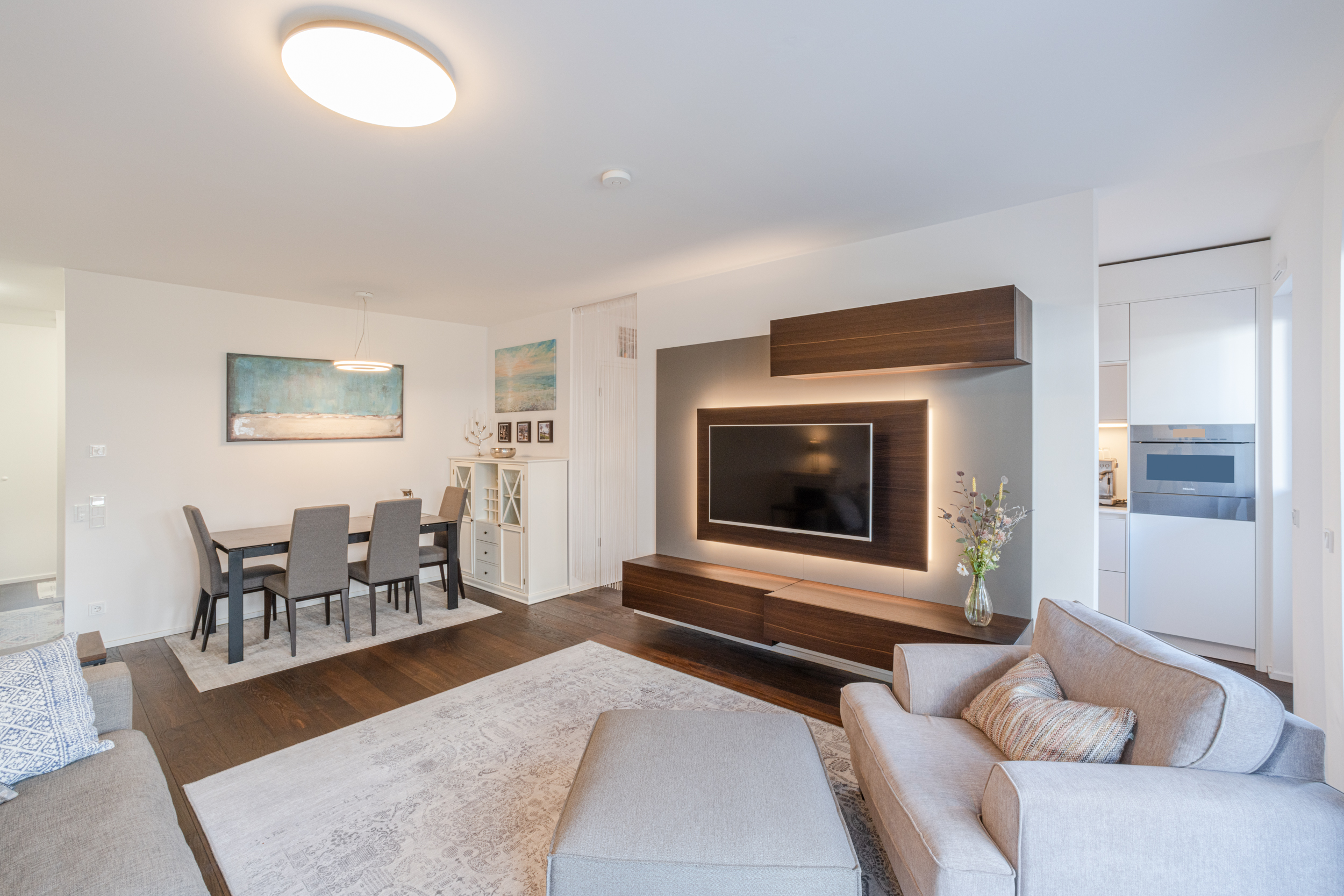 Modernes Wohnzimmer mit grauem Sofa, Esstisch, Teppich und beleuchtetem Fernseher an der Wand.