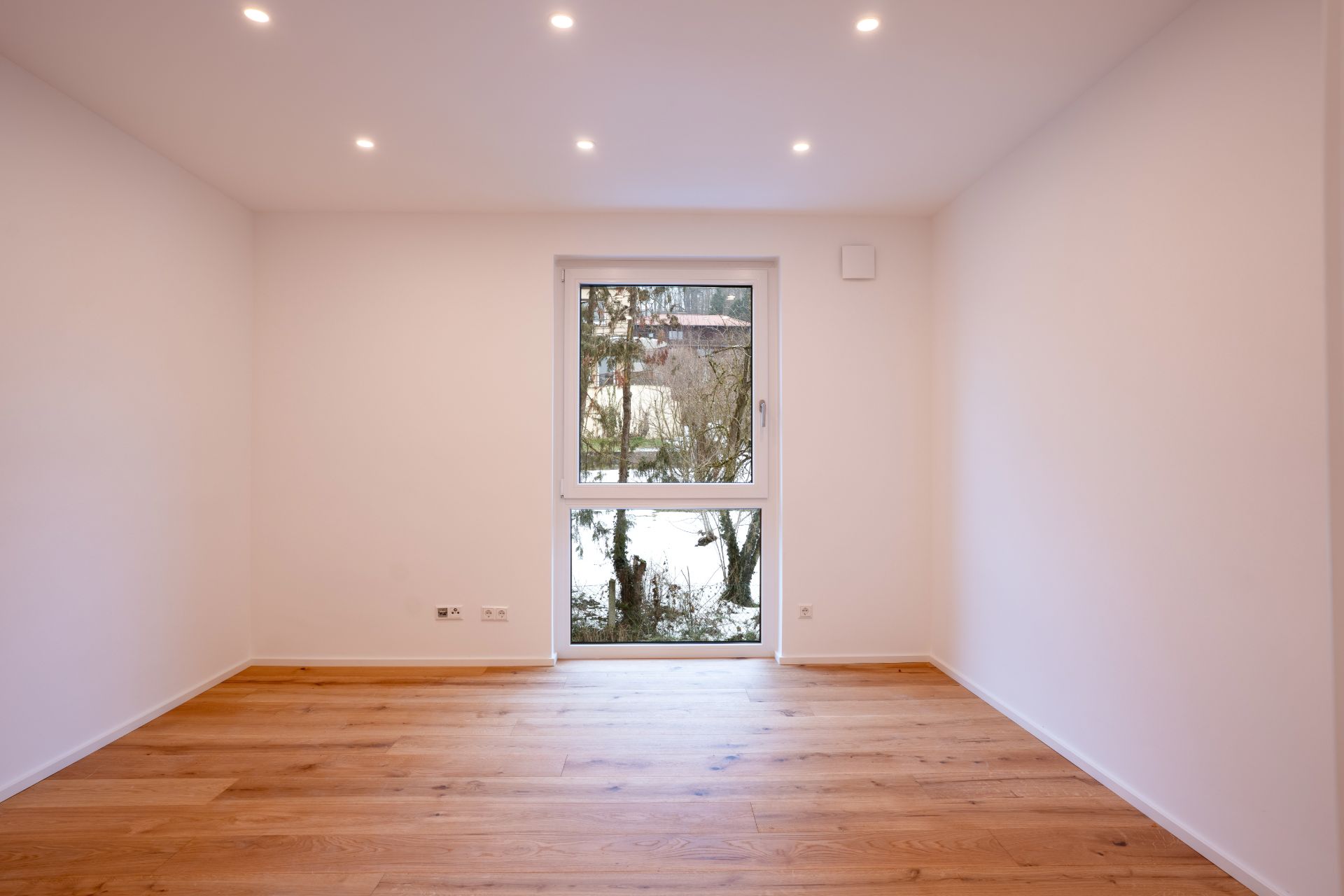 Leerer Raum mit Holzfußboden, weißen Wänden und Fenster zur Außenseite.