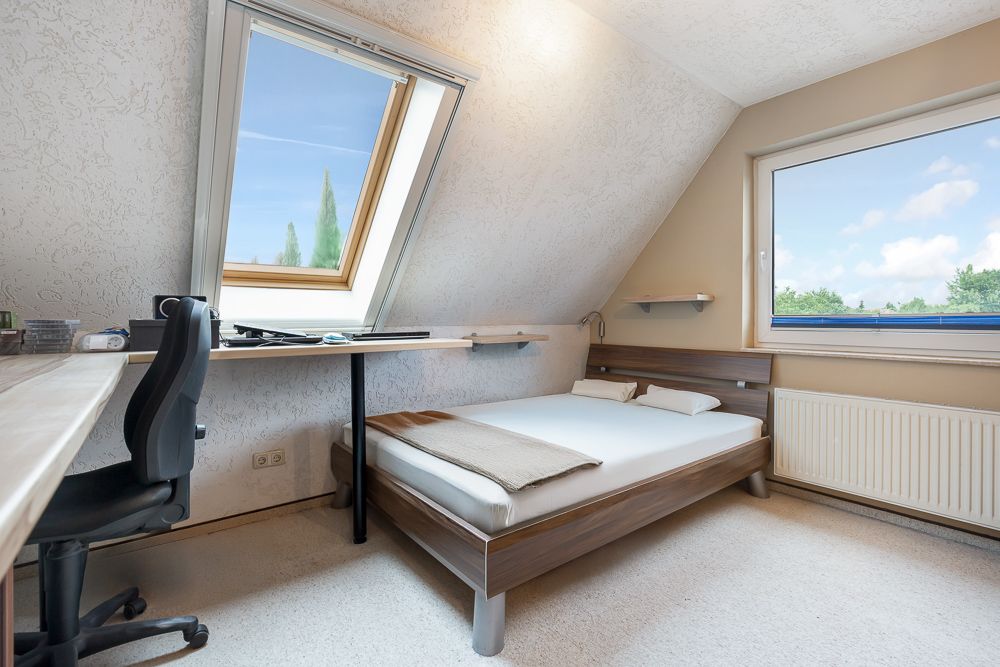 Helles Schlafzimmer mit Bett, Schreibtisch, Bürostuhl und zwei Fenstern unter einer Dachschräge.